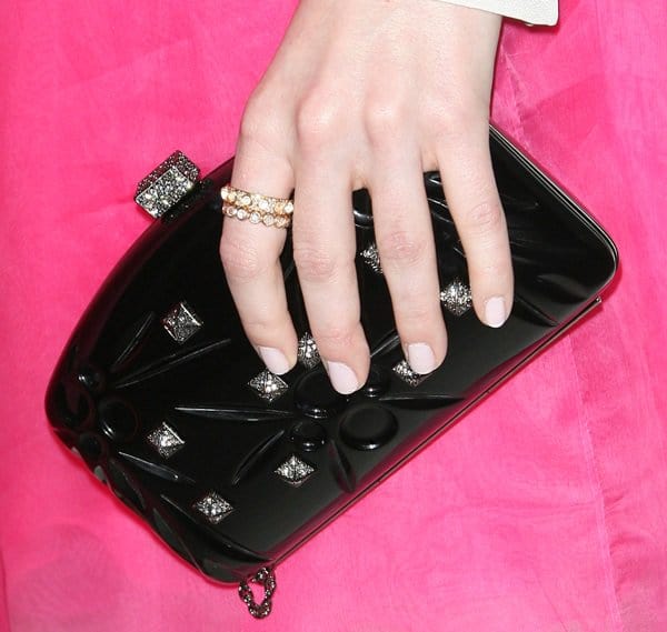 Emma Stone's accessories perfectly complement her pink collared Valentino dress, featuring a stunning black clutch adorned with sparkling details and a delicate ring on her manicured hand
