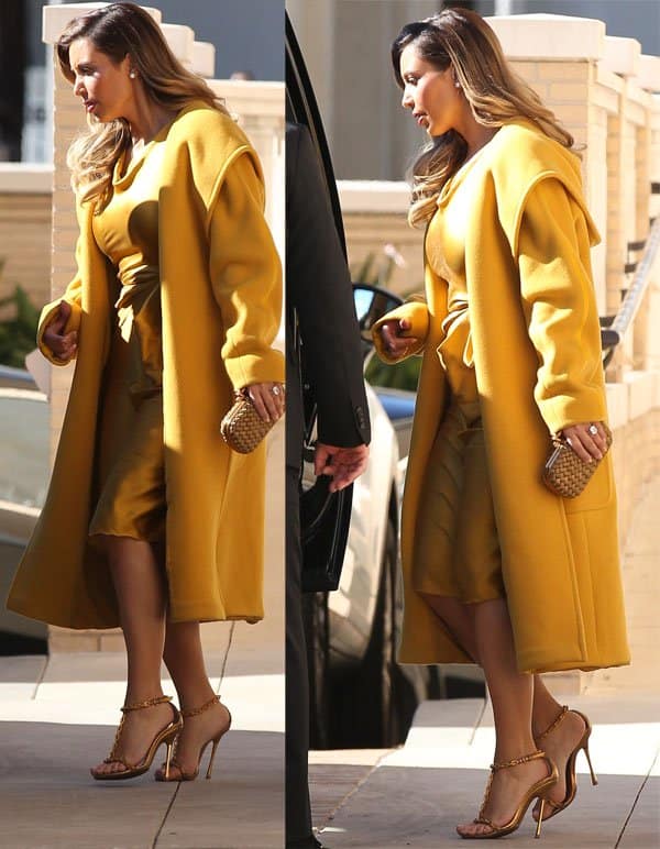 Kim Kardashian exudes confidence and style, posing gracefully in a mustard yellow silk dress paired with a matching coat and chic chain-link heels, accentuated by her elegant gold clutch