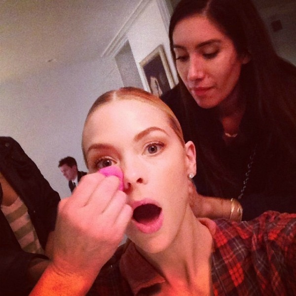 Jaime King makes a surprised expression as she gets her makeup done by a makeup artist, who is applying product to her cheek with a pink sponge, while another person looks on from behind