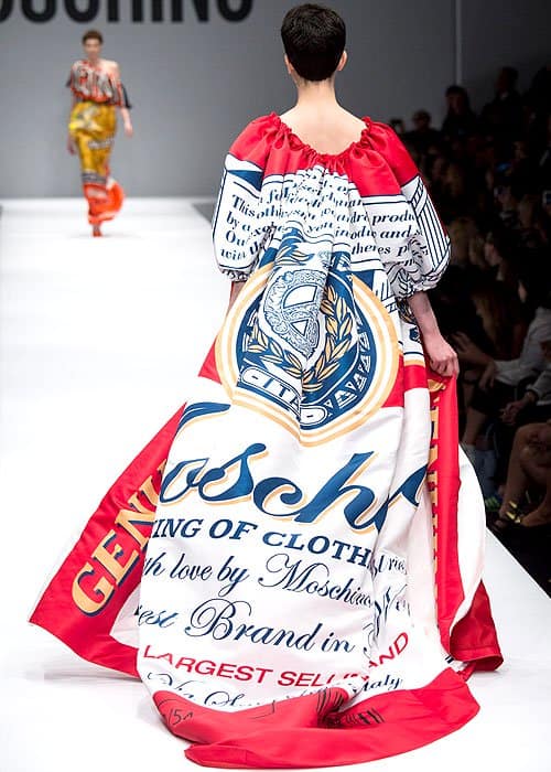 This Moschino Fall 2014 runway look showcases a model in a short, graphic-printed dress and a dramatic, flowing red and white cape, both featuring bold beer branding motifs