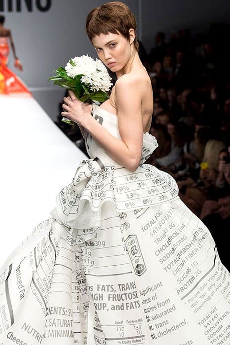 A close-up view of a model at Moschino Fall 2014 showcasing a strapless gown intricately printed with nutrition facts, featuring a ruffled waistline, as she holds a white floral bouquet