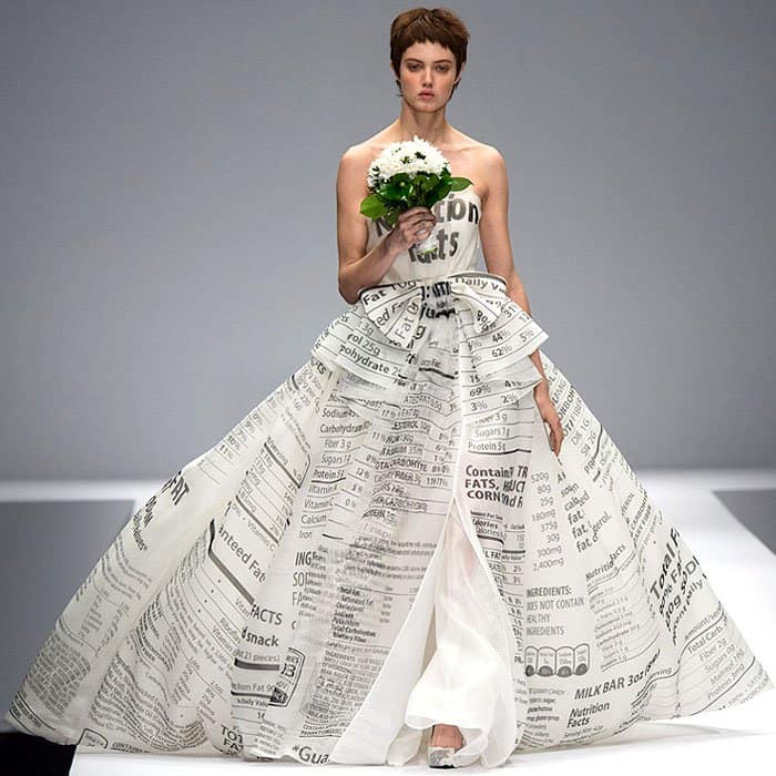 Moschino Fall 2014 showcases an elegant, strapless gown intricately printed with nutrition facts, featuring a voluminous skirt and a high slit, held by a model carrying a white floral bouquet