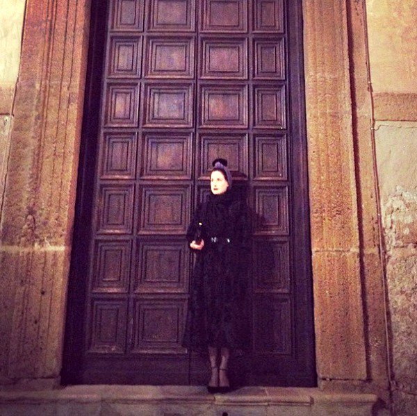 Dita Von Teese poses against a grand wooden door in Sicily, embodying timeless sophistication in a dark, chic ensemble