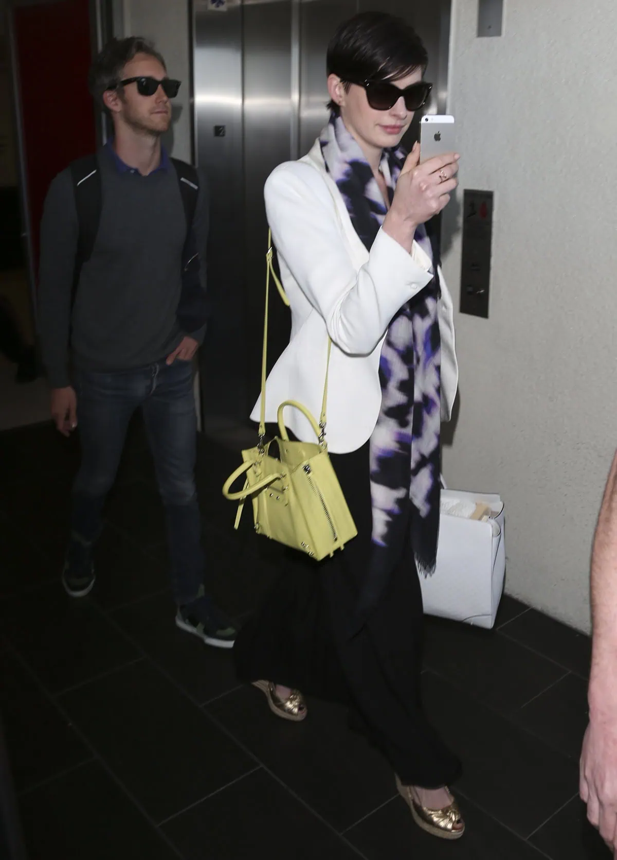 Anne Hathaway demonstrated her stylish yet witty airport demeanor in March 2014, accessorizing her white blazer and dark pants with a distinctive tie-dye scarf, a Citron Yellow Balenciaga Mini Papier A4 bag, a white Gucci tote, and Christian Dior sunglasses
