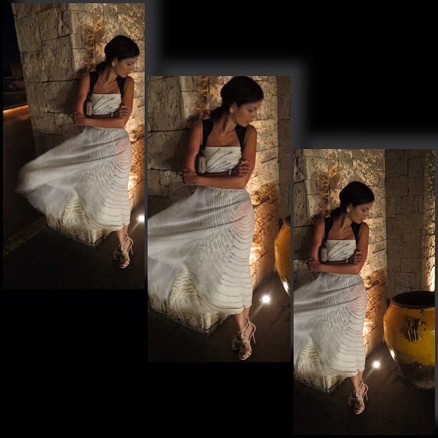 Camilla Belle leans against a stone wall in a flowing white Carolina Herrera dress, finishing the breezy evening look with delicate nude Casadei butterfly-embellished sandals that add a whimsical, feminine touch
