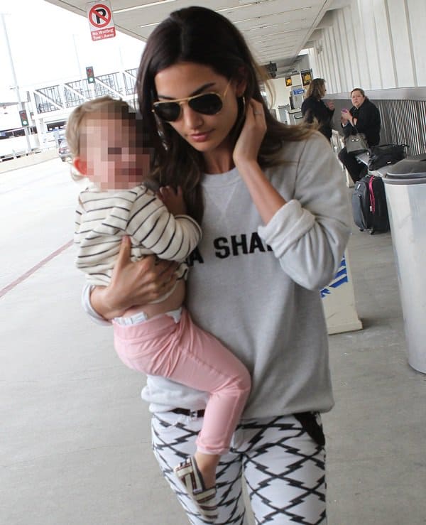 Lily Aldridge walks through LAX carrying her daughter, wearing a gray “AHA SHAKE” sweatshirt, black-and-white Ikat-print cropped jeans, and aviator sunglasses