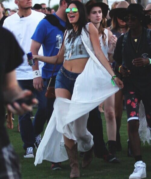 Kendall Jenner channels effortless Coachella cool in a cropped graphic tank, denim cutoffs, and suede cowboy boots, topped with a flowing white duster and mirrored aviator sunglasses