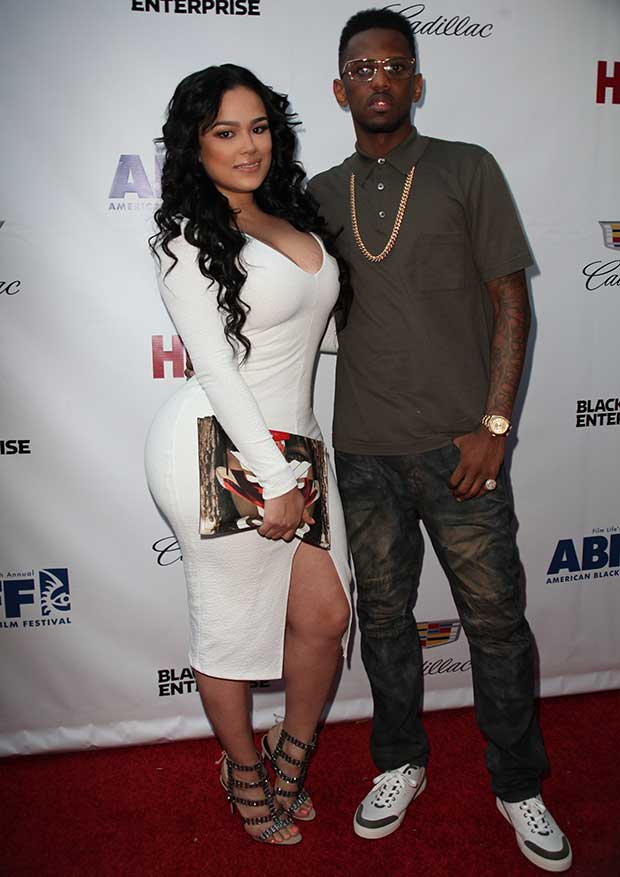 Emily B posed alongside Fabolous in a sleek white bodycon dress with long sleeves and a thigh-high slit, elevating the look with a statement Givenchy clutch and crystal-embellished Schutz sandals for a touch of red-carpet sparkle