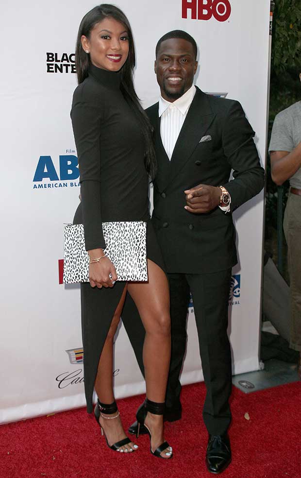 Eniko Parrish exuded modern elegance in a long-sleeved black gown featuring a daring thigh-high slit and sleek high neckline, paired with a monochrome animal-print clutch and minimalist jewelry, while Kevin Hart complemented her look in a classic double-breasted tuxedo