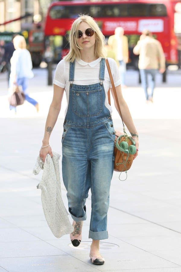 Fearne Cotton adds a feminine touch to her light-wash denim dungarees with a scalloped collar top, round sunglasses, two-tone flats, and a shoulder bag, carrying a knitted sweater for cooler weather