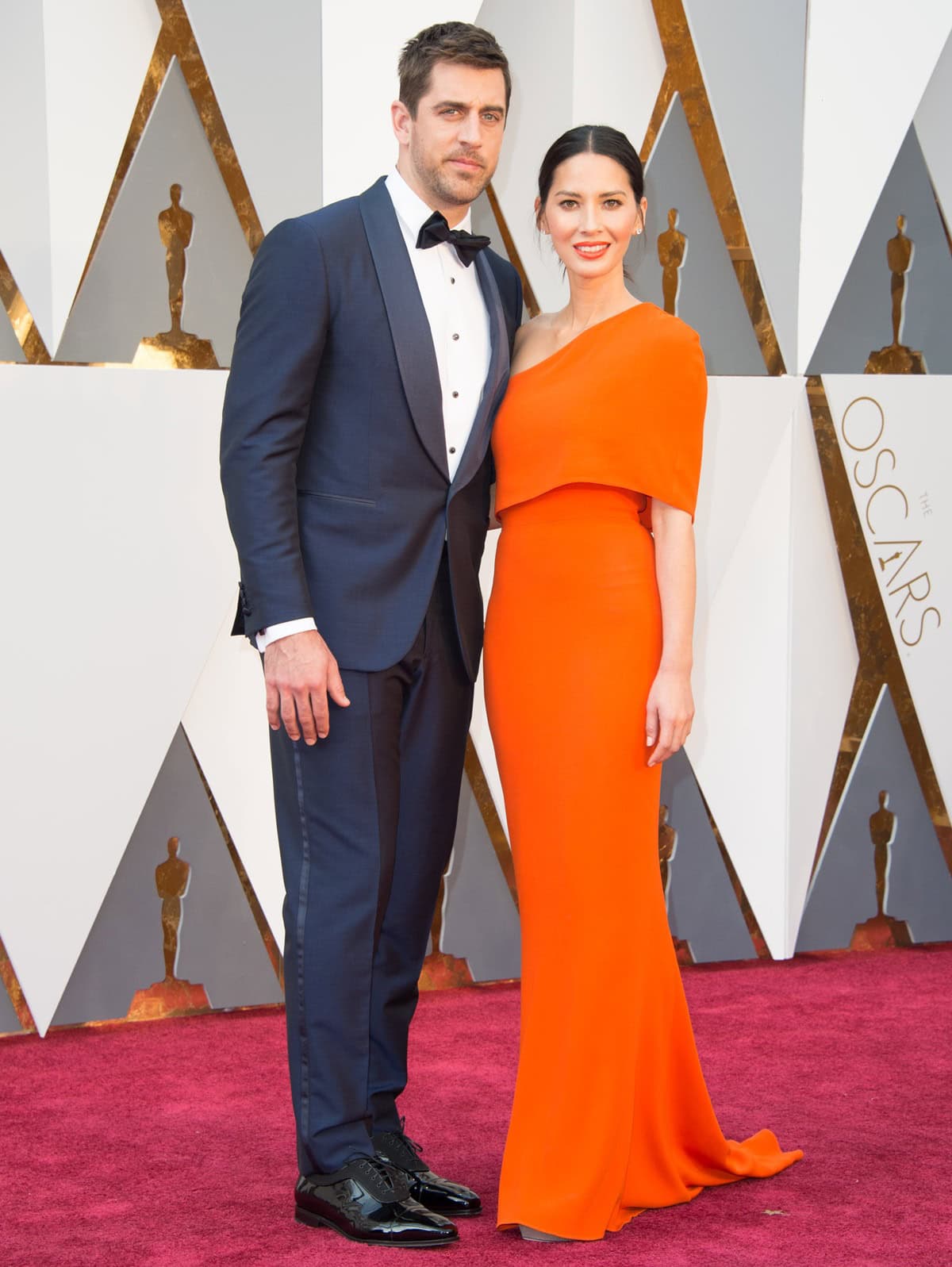 Olivia Munn brought a vivid burst of tangerine glamour to the 2016 Oscars in a sculptural Stella McCartney gown styled with diamond jewelry and silver pointed-toe heels, while Aaron Rodgers complemented her sleek red-carpet look in a classic black Isaia tuxedo with glossy Salvatore Ferragamo patent leather shoes