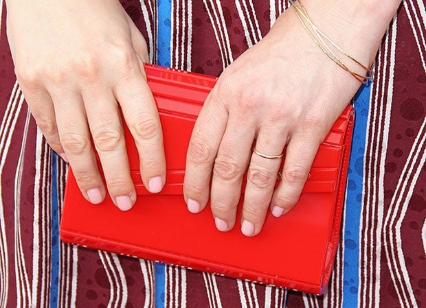 Judy Greer holds a structured red clutch while wearing delicate gold rings and thin bracelets