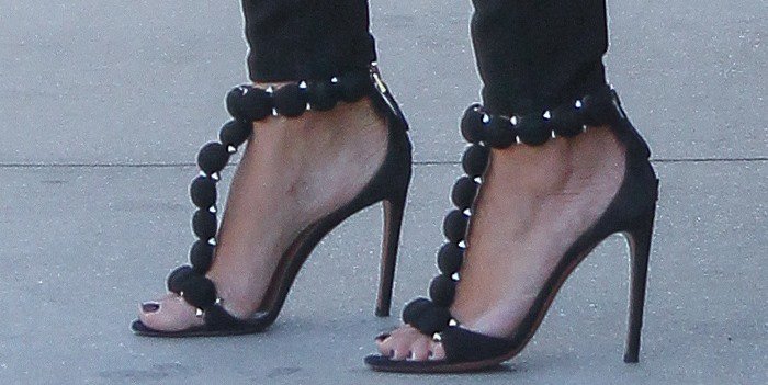 Kim Kardashian shows off her feet in Alaïa black suede studded T-strap sandals, featuring bold spherical embellishments and sky-high stiletto heels that elongated her petite 5-foot-2 frame and added an edgy contrast to her sleek, street-style look