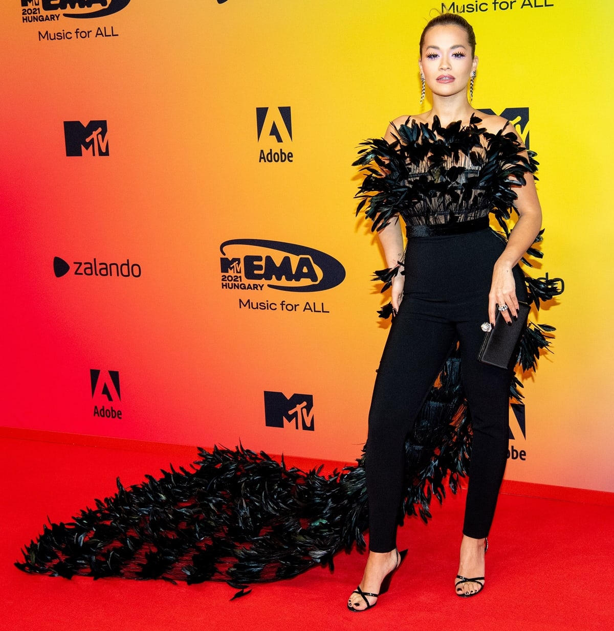 Rita Ora commands the red carpet solo at the 2021 MTV EMAs, pairing her dramatic feather-trimmed ensemble with sleek strappy Giuseppe Zanotti wedge sandals that balance the look’s theatrical edge