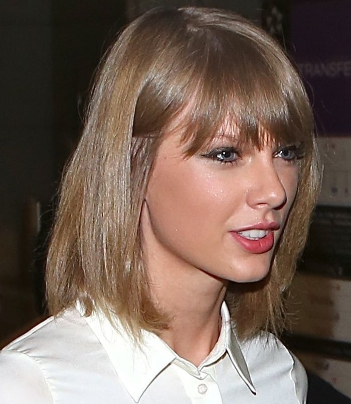 Taylor Swift is seen in close-up with a sleek shoulder-length bob and blunt bangs, wearing subtle eyeliner, rosy lipstick, and a crisp white collared shirt