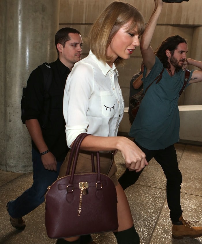 Walking through an airport terminal, she carries a structured burgundy handbag and pairs a white button-down with high-waisted brown shorts and knee-high socks