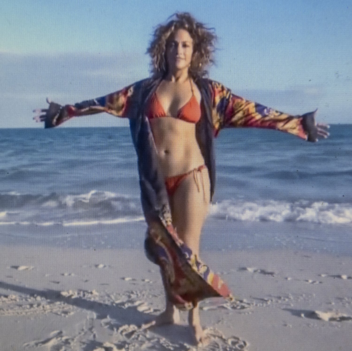 Stretching her arms toward the ocean, Jennifer Lopez channels carefree coastal glamour in a vibrant printed robe layered over a scarlet bikini