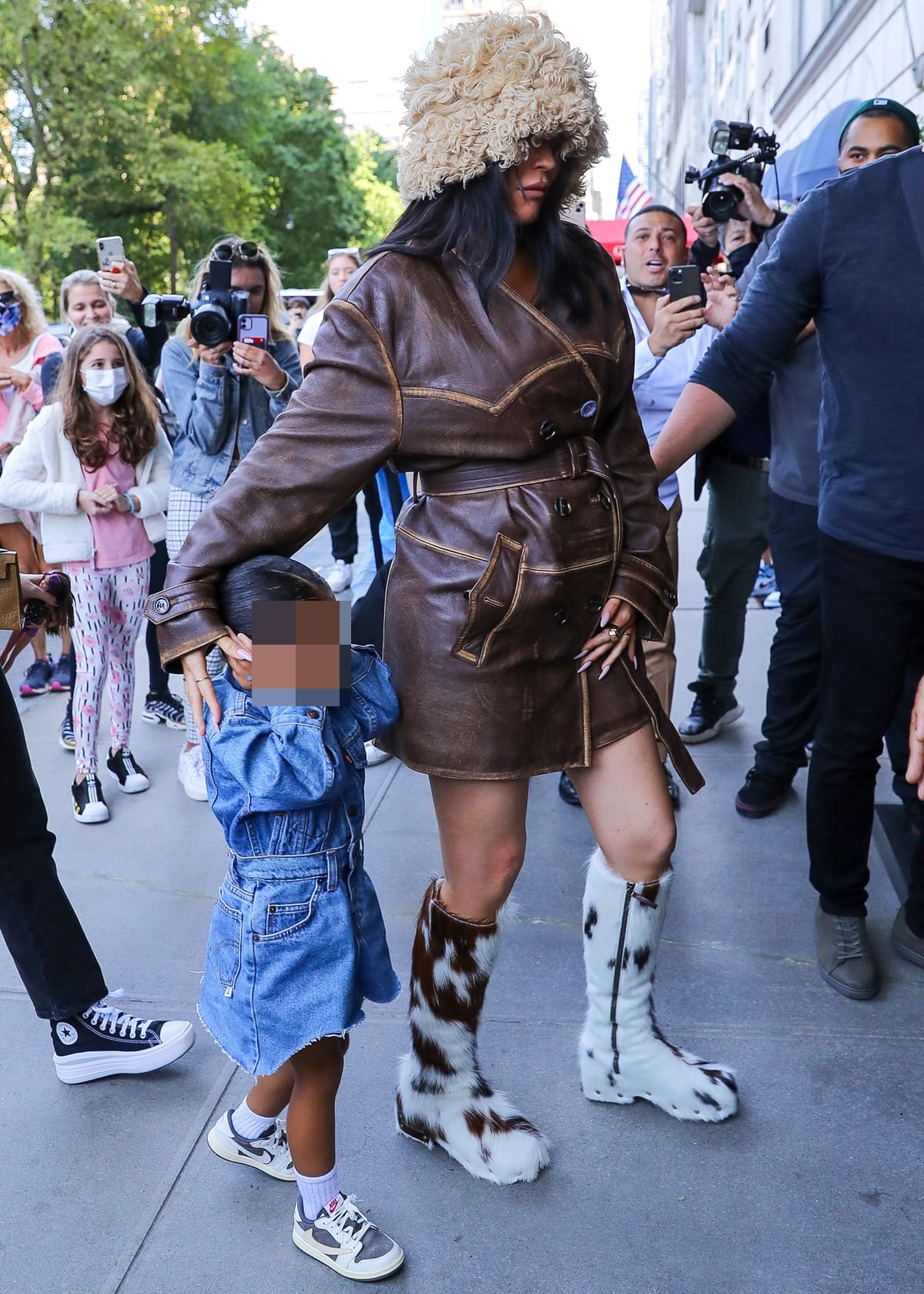 Pregnant Kylie Jenner steps out in NYC wearing a belted Acne Studios leather trench coat, shaggy bucket hat, and statement cow-print fur boots while holding daughter Stormi Webster’s hand