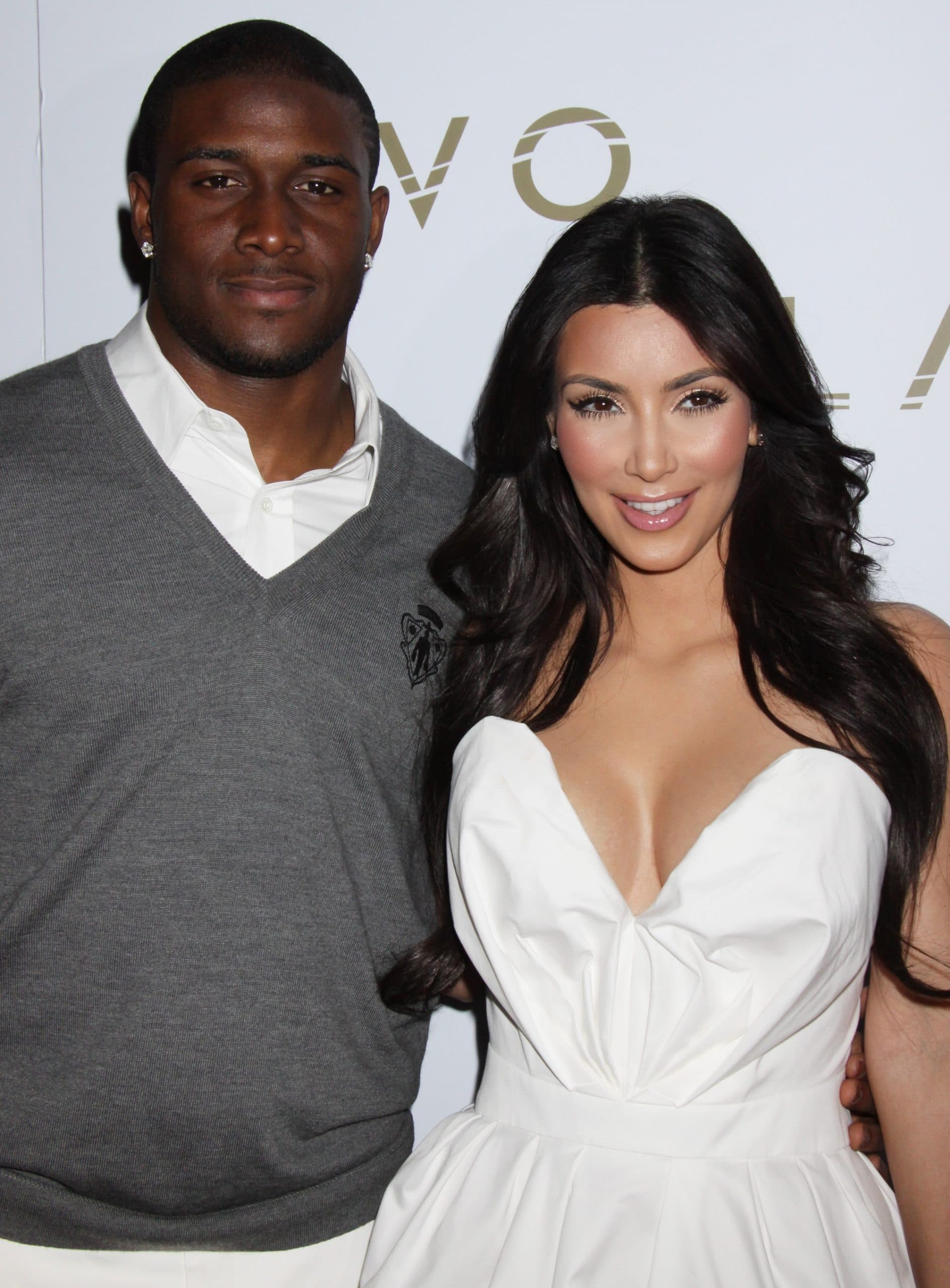 Kim Kardashian and Reggie Bush pose together on the red carpet, with Kim stunning in a strapless white dress and Reggie in a gray sweater and white shirt