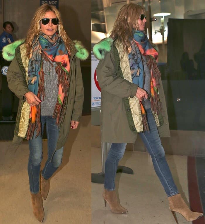 Heidi Klum wears an oversized parka with green fur trim, a colorful fringed scarf, a gray sweater, skinny jeans, and heeled cowboy ankle boots