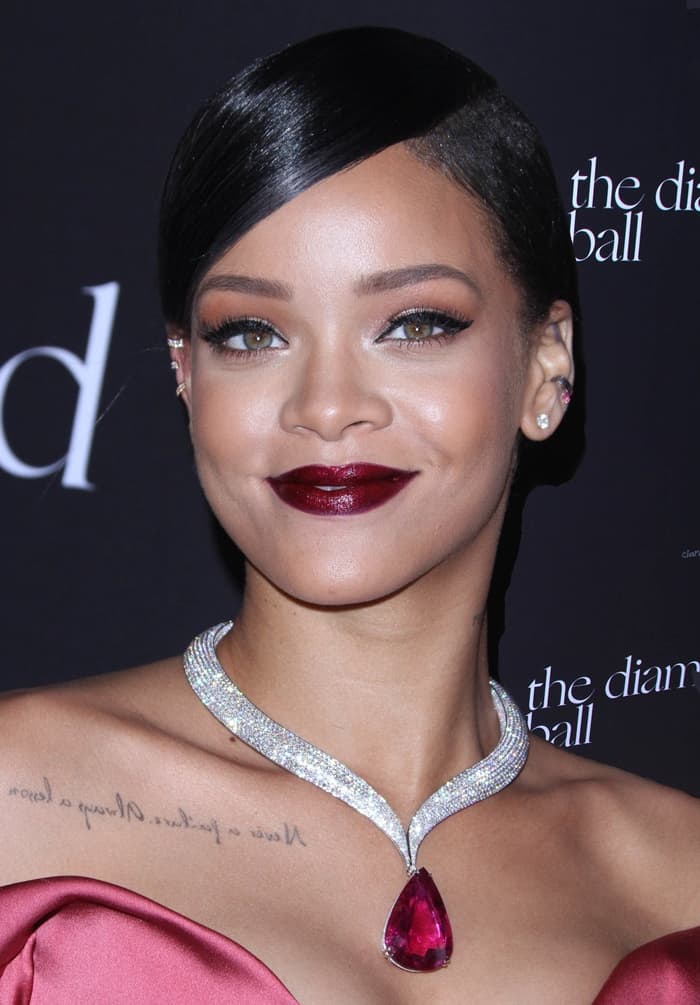 Rihanna's makeup features bold, dark berry lips, perfectly arched brows, and a subtle smoky eye with winged eyeliner that accentuates her green eyes