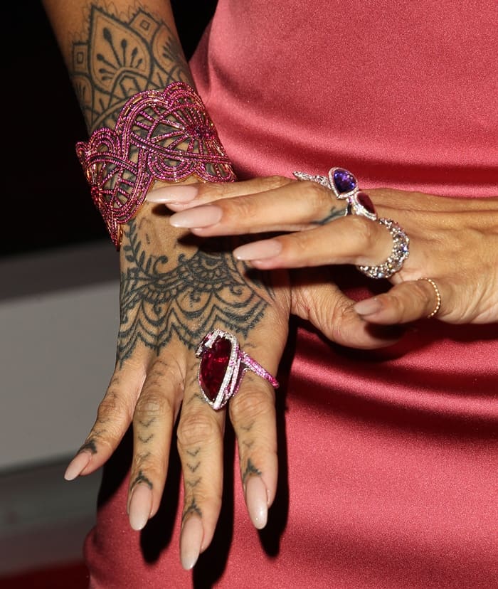 Rihanna's accessories include a striking ruby cuff bracelet, an oversized red gemstone ring, and a collection of intricate rings with colorful gemstones, all beautifully complementing her tattooed hands and elegant gown
