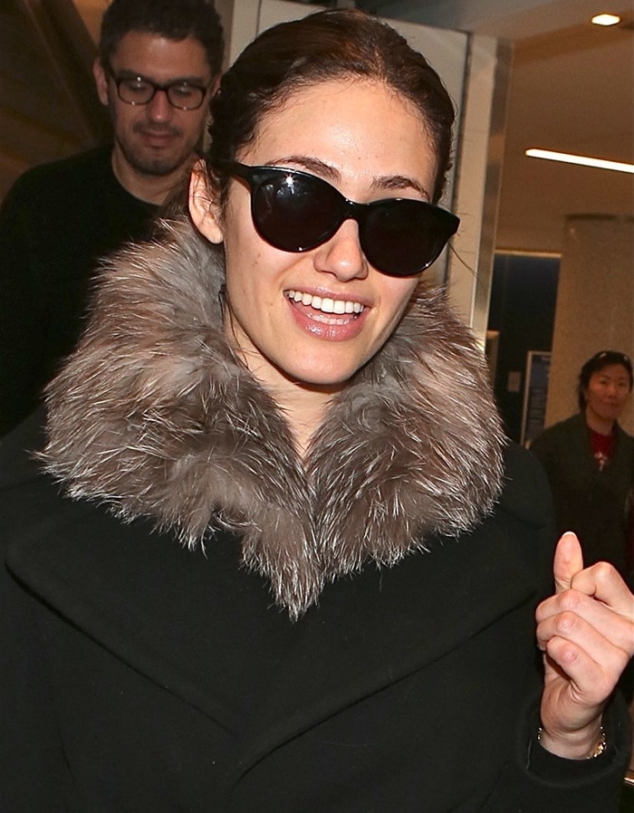 Emmy Rossum flashes a smile in a chic fur-trimmed coat and oversized sunglasses at LAX