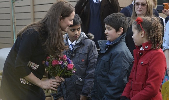 Kate Middleton smiling and receiving flowers from a young student during her school visit in a maternity shift dress