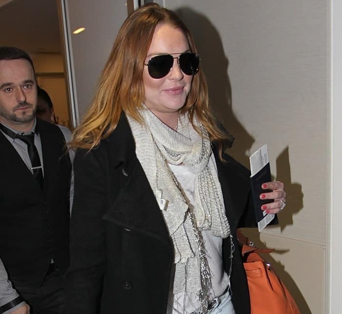 Lindsay Lohan arrives at LAX in style, wearing a chic black coat, scarf, aviator sunglasses, and carrying an orange Hermes bag