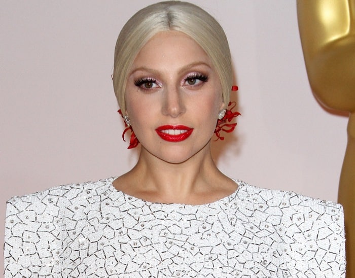 Lady Gaga styled her look with sleek platinum blonde hair adorned with red orchid blooms, bold red lipstick, and a textured white dress with structured shoulders