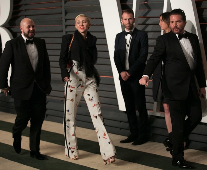 Walking alongside fellow guests, Cyrus lets the bold pop-art pattern of her trousers subtly echo the statement footwear anchoring her playful couture look