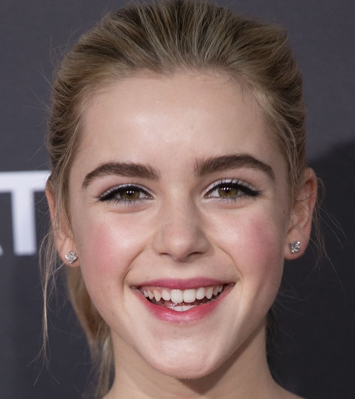 A close-up highlights Kiernan Shipka’s fresh makeup look, with softly defined eyes, natural brows, rosy cheeks, and a glossy pink lip that complements her youthful smile