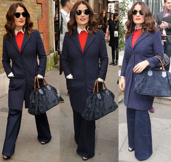 Salma Hayek strikes different poses while showcasing her retro ensemble, complete with a navy coat, flared jeans, open-toe booties, and a matching suede handbag