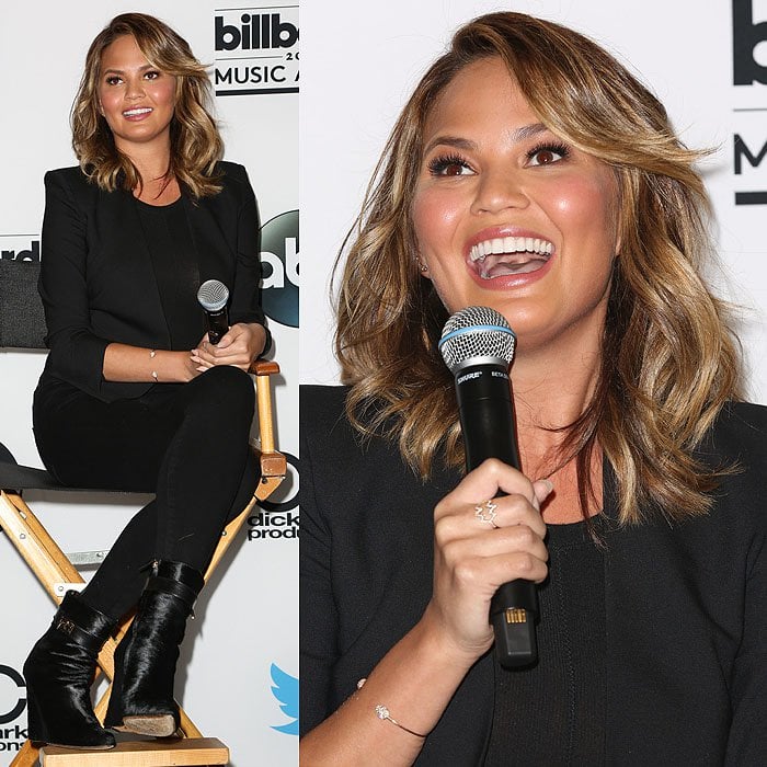 Seated onstage, Chrissy Teigen pairs skinny black jeans with sharp Givenchy sharktooth ankle boots, striking a balance between relaxed confidence and edgy sophistication