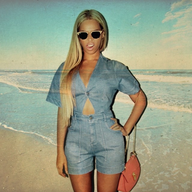With the ocean behind her, Beyoncé channels effortless summer glamour in her tailored denim romper, sleek sunglasses, and bright designer shoulder bag