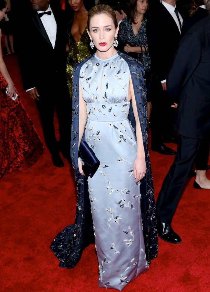 Emily Blunt exudes elegance in a custom Prada gown featuring floral embroidery and cutout details, complemented by a matching navy cape and clutch, at the 2015 Met Gala