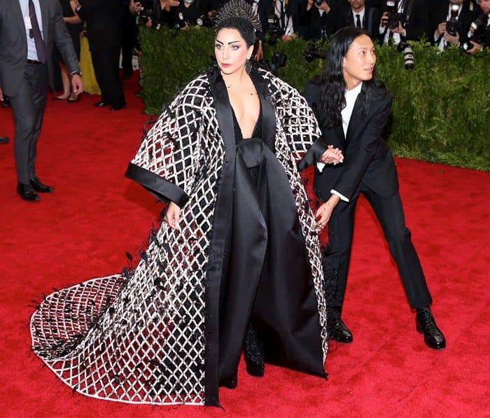 Lady Gaga commands attention in a kimono-inspired Balenciaga ensemble, featuring a dramatic lattice-patterned coat and a plunging neckline, while designer Alexander Wang adjusts her outfit on the red carpet at the 2015 Met Gala