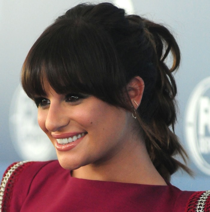 Lea Michele shows off soft glam makeup and a textured ponytail with full fringe, a romantic contrast to the strong shoulders of her embellished burgundy mini dress