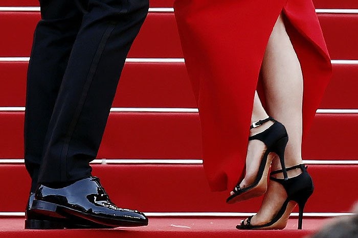 A close-up captures Benjamin Millepied's polished black dress shoes and Natalie Portman's elegant black satin sandals on the vibrant red carpet stairs
