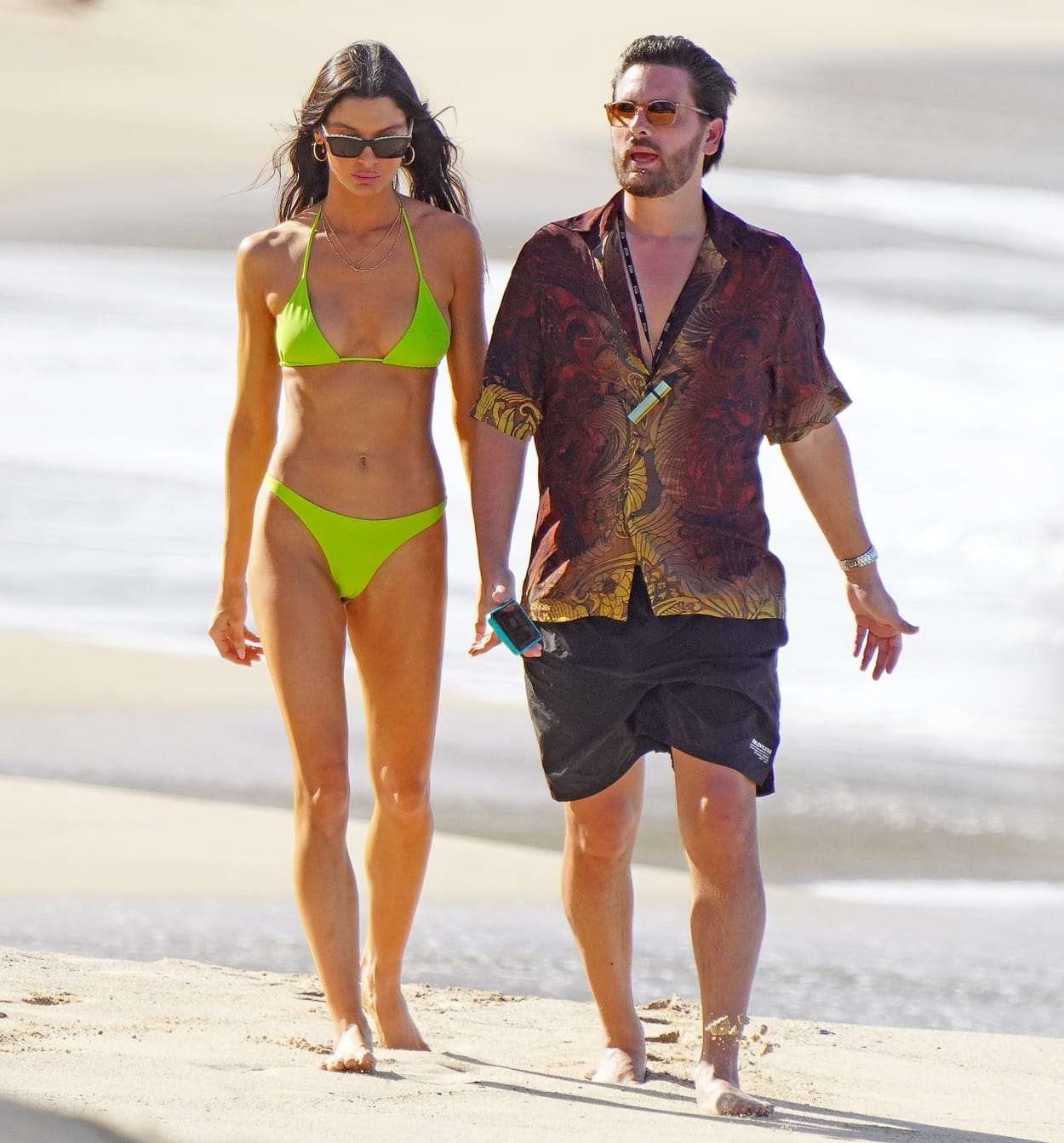 Scott Disick and his girlfriend Bella Banos walk the beach in St. Barts on December 21, 2021, with Bella stunning in a neon green bikini and Scott keeping it casual in a tropical-print shirt, black swim shorts, and sunglasses
