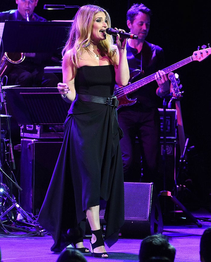 Idina Menzel stuns on stage in a strapless black belted dress paired with striking black Miu Miu suede wedge sandals featuring bold cutouts and peep toes