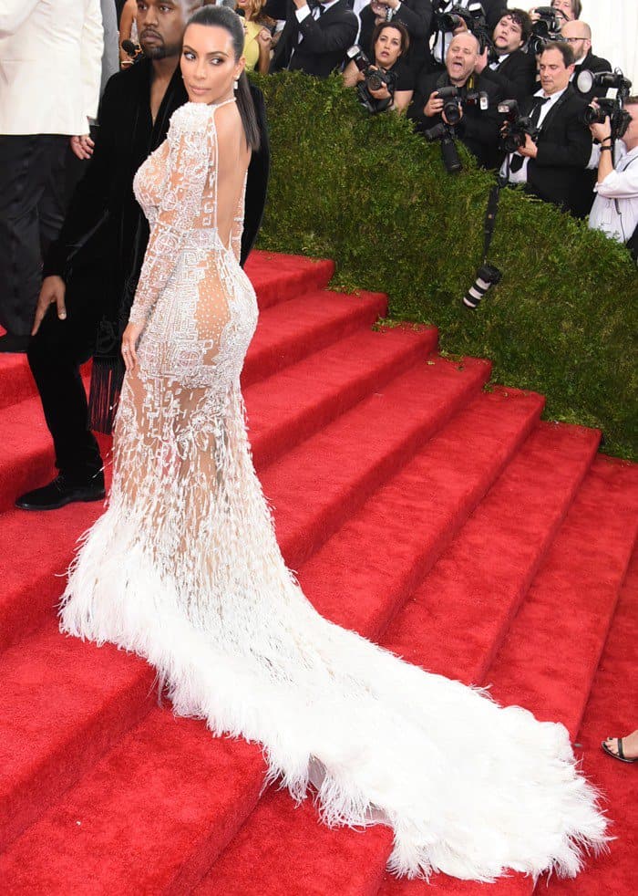 Kim Kardashian's dress at the Met Gala features a sheer white design by Peter Dundas for Roberto Cavalli, with intricate lace detailing and a long feathered train that accentuates her curves