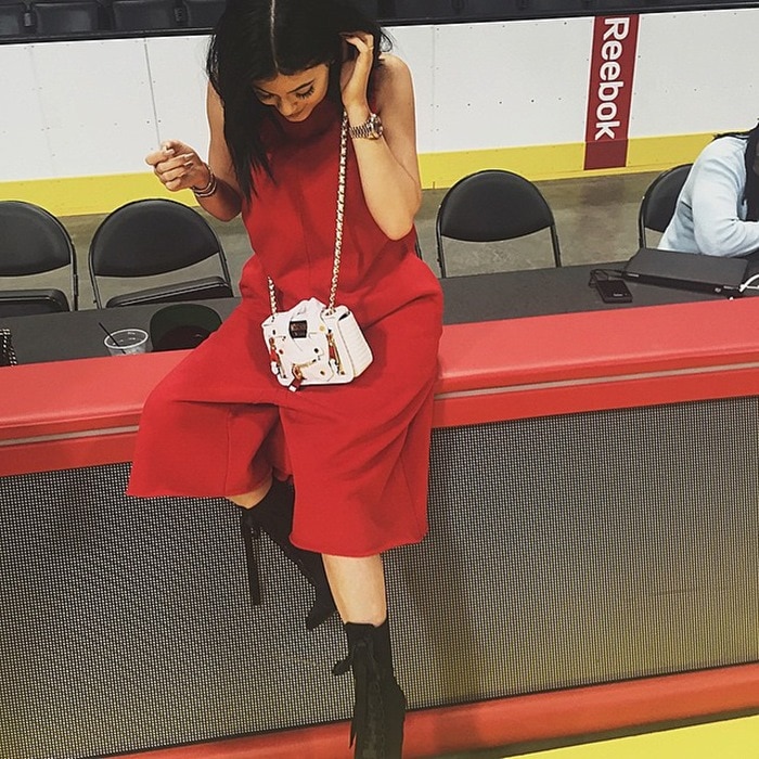 Shared on June 9, 2015, Kylie Jenner sat courtside in a bold crimson jumpsuit styled with Giuseppe Zanotti x Balmain “Uma” boots, a Moschino Smakker Biker shoulder bag, and Dior Technologic sunglasses, blending sporty edge with high-fashion polish