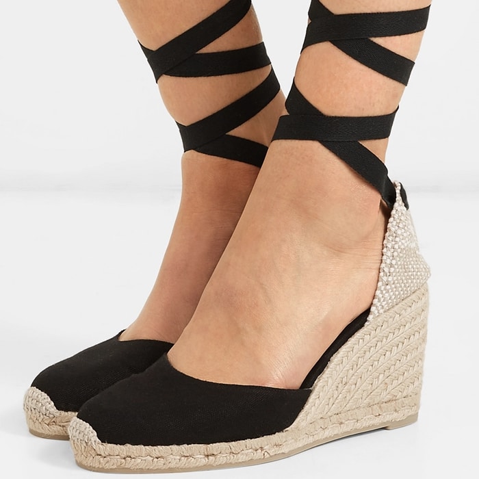 Castañer elevates summer sophistication with the black “Carina” espadrille wedges, featuring canvas uppers, wraparound ankle ties, and hand-braided jute heels that balance timeless craftsmanship with effortless elegance