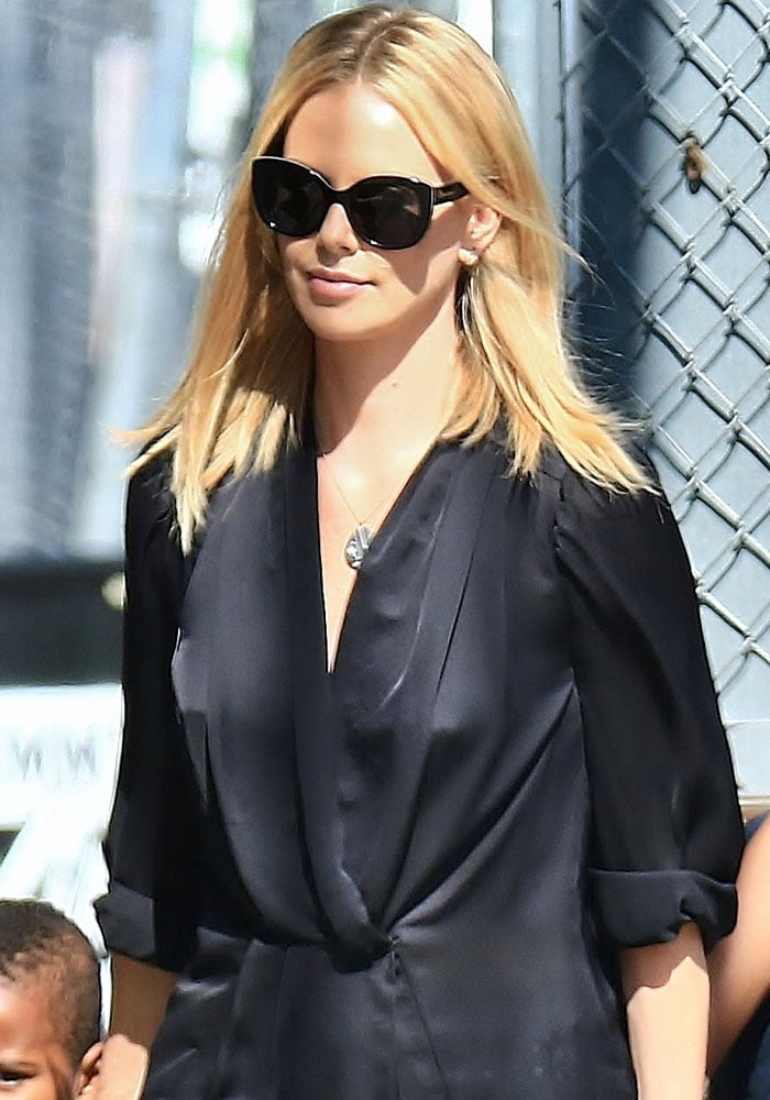 Charlize Theron, wearing dark sunglasses and a pendant necklace, is seen in a close-up shot of her oversized, tie-front black blouse