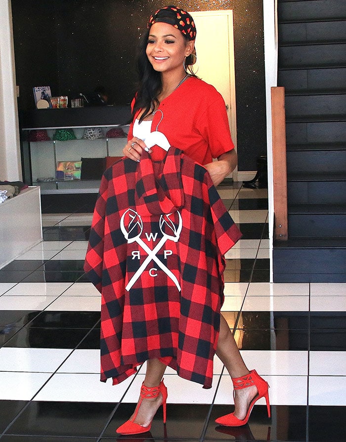 Christina Milian showcases a bold red and black outfit, featuring a red t-shirt, a white top underneath, a black and red plaid hoodie from her "We Are Pop Culture" brand, and striking red ankle-strap heels, perfectly blending streetwear with elevated footwear