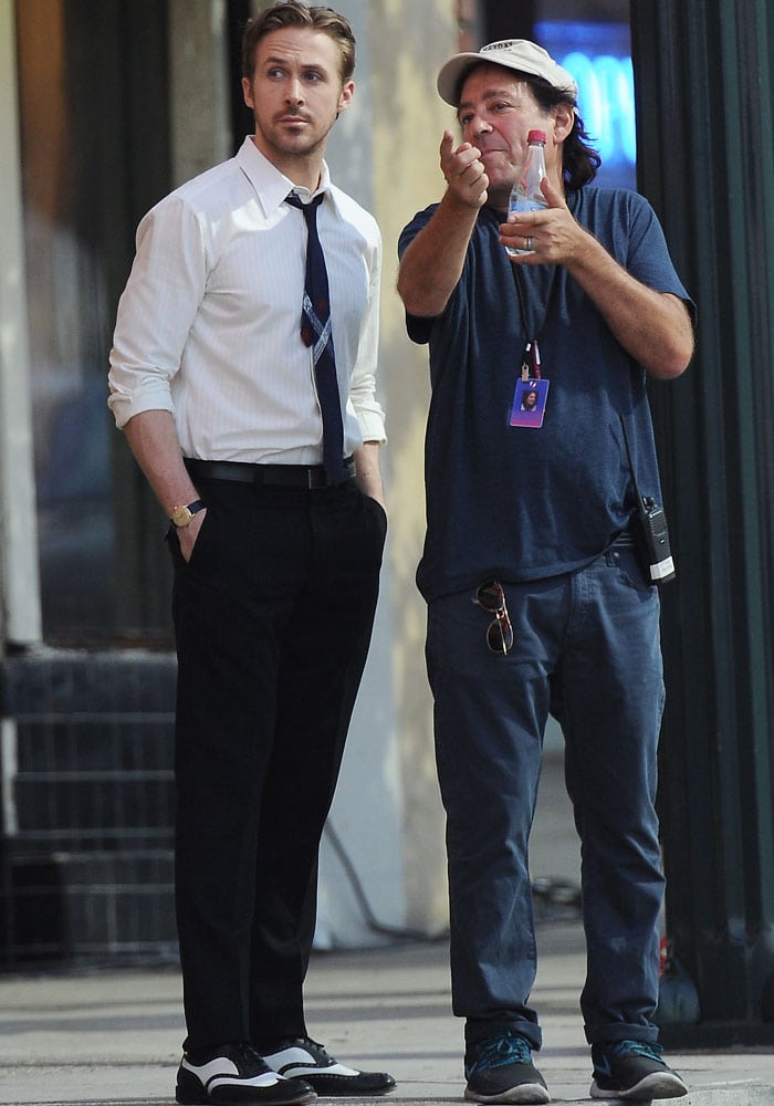 Ryan Gosling receives last-minute direction on set while dressed in character, wearing a crisp white dress shirt, dark slacks, a patterned tie, and black-and-white spectator shoes that nod to vintage jazz style