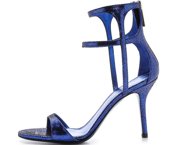 Tamara Mellon’s “Glow” sandals dazzle in crackled metallic blue suede, featuring sleek ankle straps and slender stiletto heels that deliver a bold pop of color with elegant evening appeal