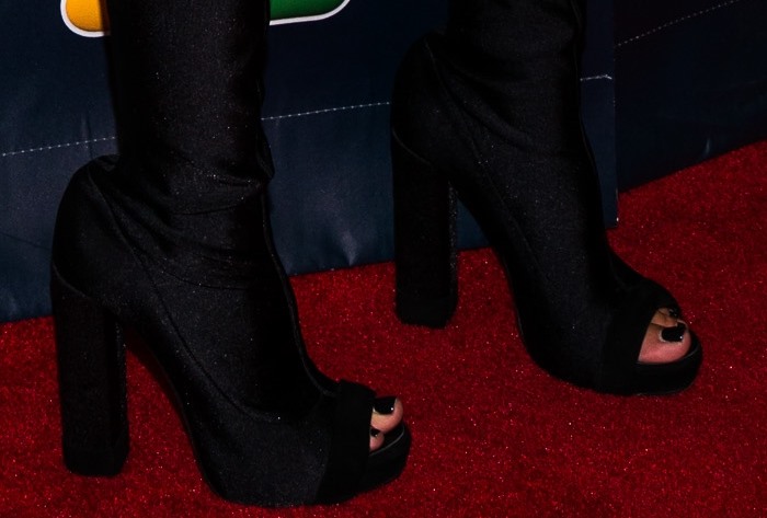 Her matching peep-toe platform boots—crafted in a satin-like finish with towering block heels—extend the outfit into a continuous line, blurring the boundary between garment and footwear