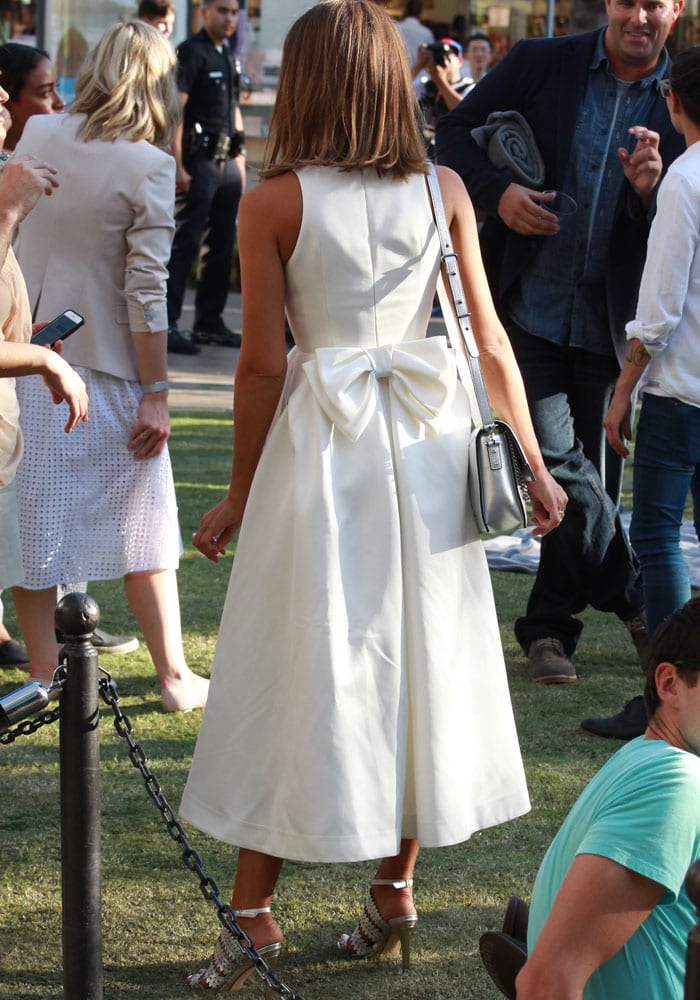 Jessica Alba turns to reveal the oversized bow detail on the back of her elegant white midi dress, styled with metallic Eight Fifteen “Bonfire” heels and a sleek silver crossbody bag at her Honest Beauty launch event
